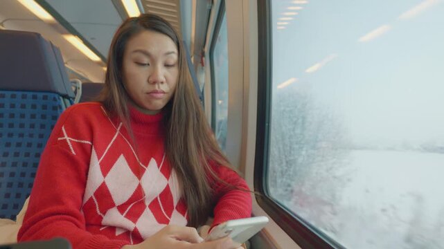 A young person in a cozy red sweater is focused on their smartphone while traveling on a train. The snowy landscape outside the window adds a serene backdrop to the commuting experience.
