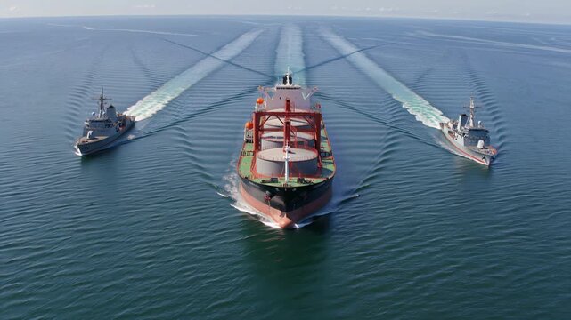 Cargo ship escorted by two naval destroyers across open ocean. Commercial maritime transport with military protection. Aerial wide shot.