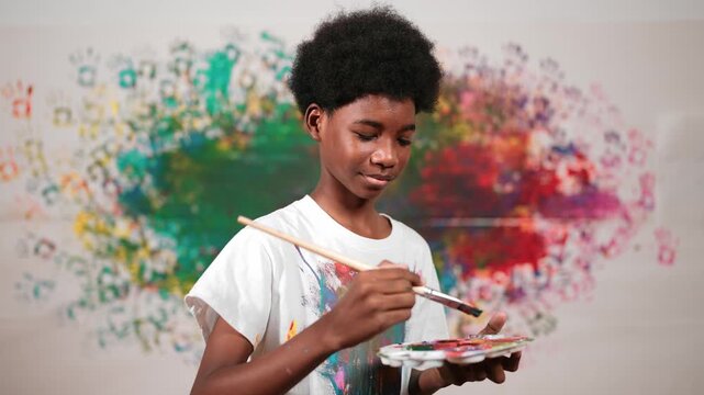 African boy standing at colorful stained wall while holding brush and point at camera. Happy high school student wearing white shirt with stain color and look at camera with confident. Edification