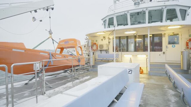 A ferry deck adorned with ice and snow, showcasing an orange rescue boat and a chilly atmosphere. The scene captures the essence of winter travel in a serene marine environment.