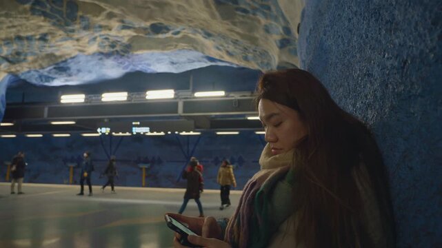 A contemporary underground station setting featuring travelers engaged with their mobile devices. The space is characterized by unique architectural elements and vibrant lighting that enhances the com