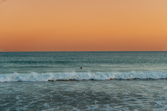 Atlantic Ocean coastline with crashing waves rolling back into the sea, blue water, and a glowing sunset creating a dramatic coastal seascape and man in the ocean.
