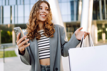 Smiling woman, holding phone in hand, stands in front of modern shopping center with shopping bags. Young woman enjoys sunny day after online shopping. Concepts of shopping, consumerism. © maxbelchenko