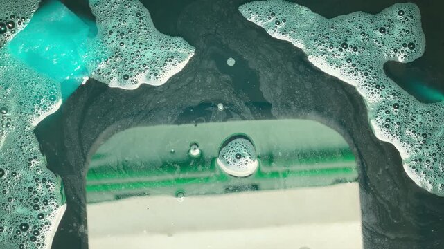 Bubbles and dark blue liquid mix inside a public restroom toilet. The swirling foam creates a cold, clinical atmosphere. It feels sterile but evokes a sense of unease and industrial grit.
