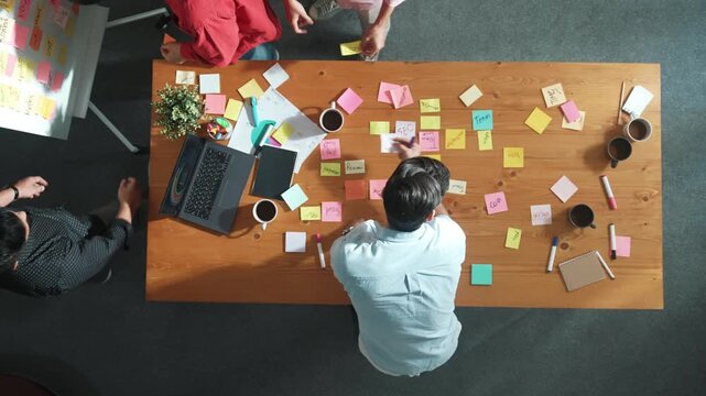 Top view of skilled business people discuss plan at meeting room while writing idea at sticky notes and stick at whiteboard. Group of diverse team working together to planning strategy. Convocation.
