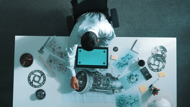 Top view of skilled engineer writing in paper while looking at tablet display green screen with turbine engine sketch design and metal gear and architectural equipment scatter around. Alimentation.