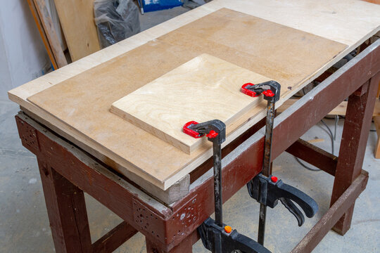 Woodworking clamp holding plywood piece on a workbench. Carpentry and furniture making process. Manual labor tool for fixing material during construction or diy renovation project