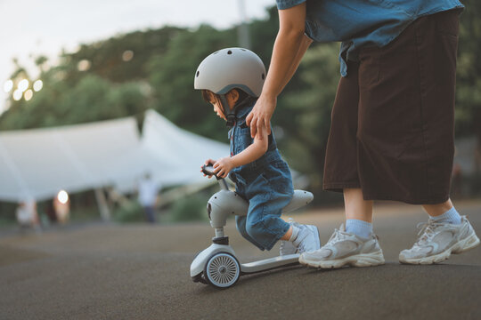 Side view of cute little Asian girl wearing helmet riding scooter balance bike at public park during evening mother teaching and supporting toddler active childhood vacation and family lifestyle.