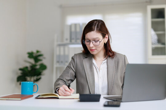 Young professional businesswoman writing notes in a notebook, managing tasks, and working on her laptop in a contemporary office