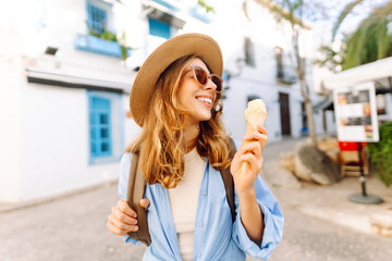 Fototapeta premium A happy tourist in a hat, holding an ice cream cone, strolls along the sunny streets of the city. The young woman admires the city's architecture and eats ice cream. Concept: tourism, vacation.