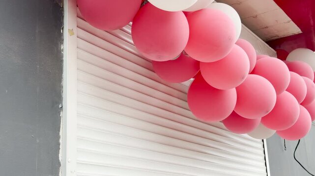 A bunch of pink and white balloons hangs over a closed metal roller shutter on a gray wall. The busy street outside generates a continuous, distant rumble of city traffic.
