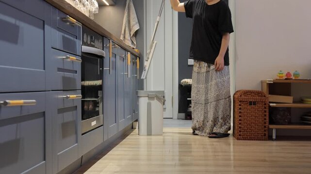 A teenager mops a light wooden floor beside blue kitchen cabinets during a home cleaning routine.