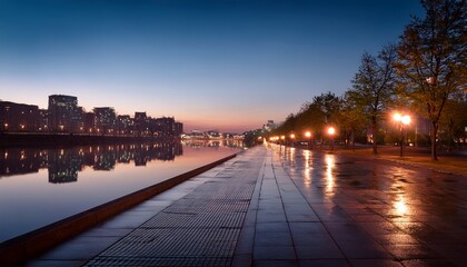 Fototapeta premium wet concrete walkway beside a calm river reflects streetlights at dusk creating a serene urban landscape