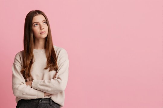 Young woman with arms crossed looks up thoughtfully. Female is in studio against pink background. She appears to be contemplating future plans or goals with a pensive expression.