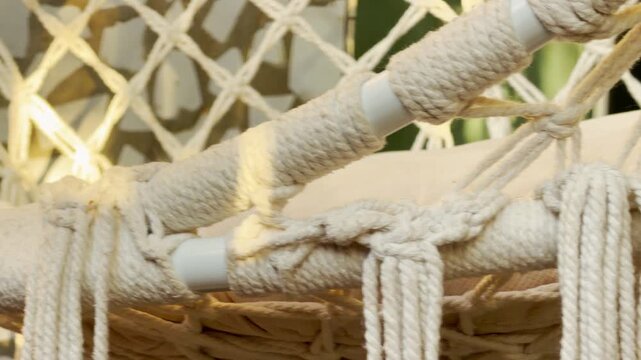 A handmade macrame swing chair hangs peacefully in a sunlit outdoor space. The warm light hitting the woven ropes creates a cozy and relaxing feeling. It looks like a perfect spot for a quiet rest.