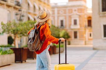 A young tourist with a yellow suitcase and backpack walks through a sunny city square. A woman in a hat enjoys her travels. Concepts of tourism, vacation, and relaxation. © maxbelchenko