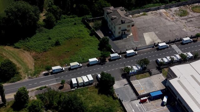 Logistics Convoy: An aerial view captures a long line of white vans and trucks forming a logistical convoy on a rural road, heading towards an industrial area with buildings and greenery surrounding.
