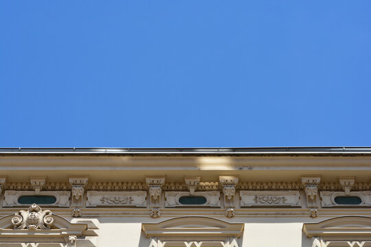 Low angle view of a decorative historic building facade with detailed plaster stucco ornaments and a roof cornice under a clear blue sky.