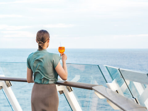 Fashionable woman with a glass of drink standing on the empty deck of a cruise ship against the backdrop of sea waves. Sunny day. Chill lifestyle, luxury travel. Perfect for holiday and travel content