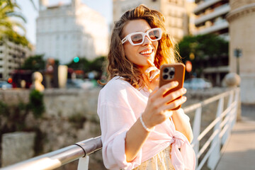 Obraz premium A young woman, holding a phone, strolls through a sunny city near a park. A beautiful woman enjoys the sunset in a stylish dress and with a smartphone. Concept of fashion, style, and relaxation.