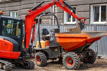 Red mini excavator with its arm raised working next to a small orange site dumper on an urban construction site by an old building, metal fence and dirt in daylight. © Adam
