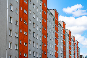 Long multi-story residential apartment block with a repetitive pattern of windows on a grey and orange facade, shot at an angle in daylight with blue sky and white clouds. © Adam