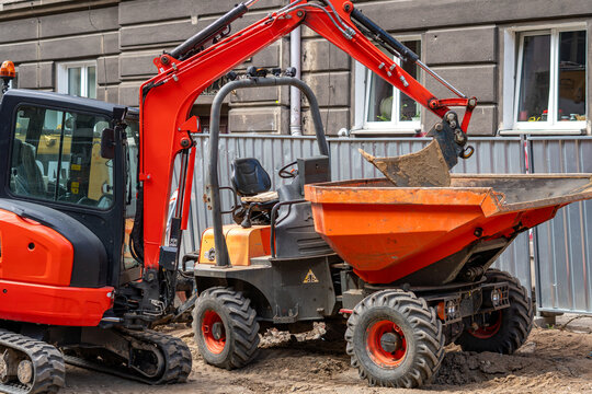 Red mini excavator with its arm raised working next to a small orange site dumper on an urban construction site by an old building, metal fence and dirt in daylight.