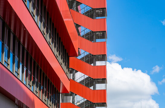 Modern building facade made of red corrugated metal sheet and an outdoor zigzag emergency staircase shot from a low angle, glass windows reflecting light, blue sky with white clouds.
