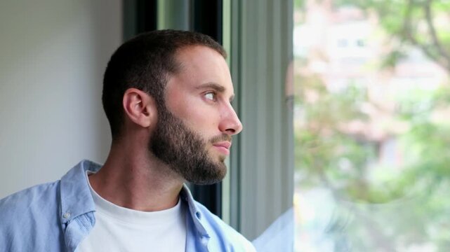 Thoughtful young man with a beard looking outside through the window, reflecting on life with a pensive expression on his face while contemplating his future from his apartment