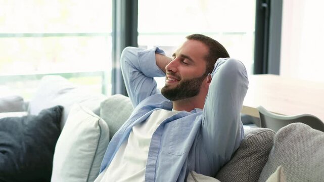 Handsome serene young man relaxing on a comfortable sofa at home, leaning back with hands behind head, enjoying a peaceful moment of tranquility and satisfaction in his living room