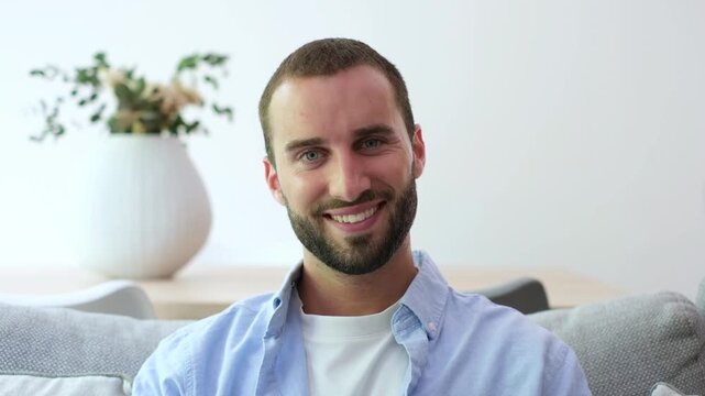 Handsome caucasian man with a beard and blue eyes sitting on a sofa in a modern living room, looking at the camera and smiling, showing confidence and a positive attitude
