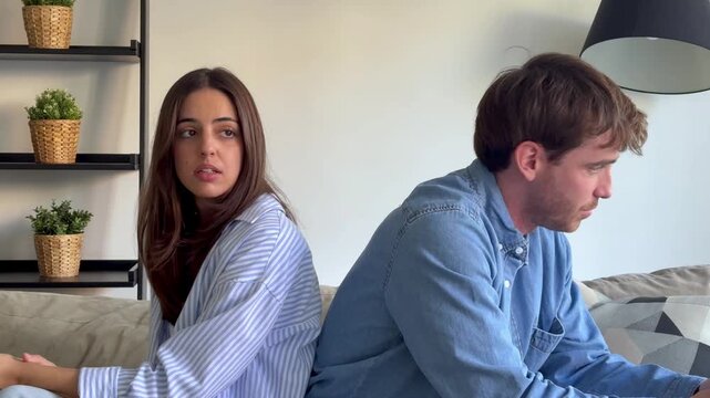 Young frustrated couple sitting back-to-back on the sofa, arguing about their relationship problems and gesturing angrily, showing conflict, misunderstanding, and emotional distress