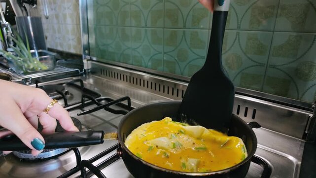 Close up of a person's hands using a spatula to fold a delicious, fluffy omelette with fresh green onions in a frying pan on a gas stove, preparing a healthy and protein rich morning meal