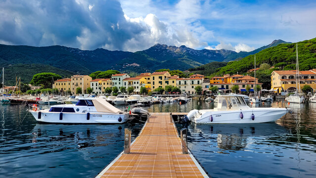 Marciana Marina, Elba, Italy - September 21, 2024. Panoramic view of the Mediterranean Riviera and a wooden path leads to yachts. In the background are typical Tuscan houses.