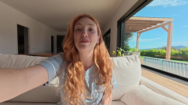 Beautiful redhead woman in pajamas speaking to the camera, vlogging from her bright, modern living room with a view of the sunny garden and patio on a beautiful summer morning
