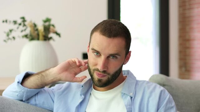 Charming handsome young man with a beard and blue eyes sitting on a sofa, looking at the camera, winking, and smiling seductively with a confident expression in his modern living room