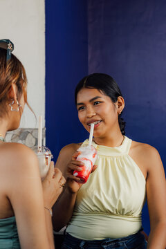 Alegre joven hispana con brackets tomando un batido de fresa y celebrando el D&iacute;a de la Juventud con su amiga en una cafeter&iacute;a.