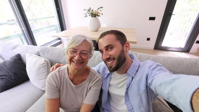 Young adult son holding his phone and taking a happy selfie with his elderly mother while sitting together on the sofa in their modern living room, showing intergenerational love and connection