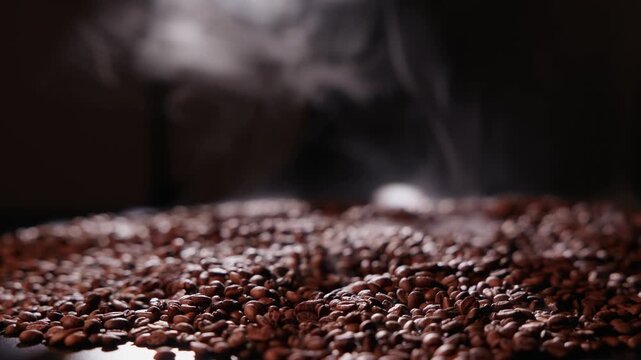 Slow motion steaming coffee beans piled on dark tabletop studio closeup with rising smoke and shallow depth.