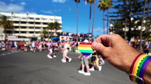 Close-up of a hand wearing a rainbow wristband and holding a heart-shaped rainbow symbol, with a blurred gay pride parade and a festive crowd marching in the background on a sunny day