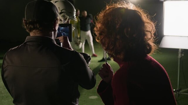 Medium arc backstage shot of American football sportsman in uniform jersey and helmet praying before match, cameraman filming repeat takes and female producer directing shooting production work