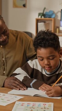 Medium vertical shot of supportive mature African American father encouraging little son during handwriting practice, gently patting on head, while helping with elementary school homework