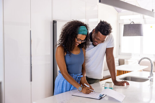 Couple standing at kitchen island, woman signing clipboard, pen in hand while man leaning, smiling