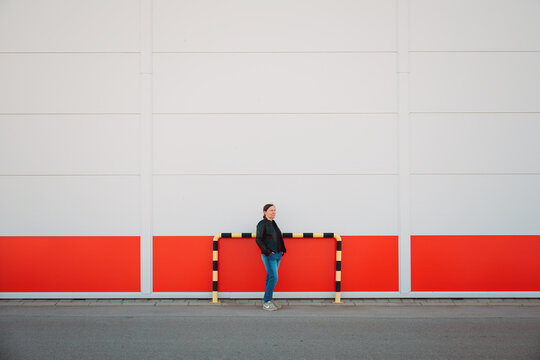 A casually dressed woman stands against a bold red and white industrial wall, conveying urban minimalism, contrast, solitude and contemporary street style.