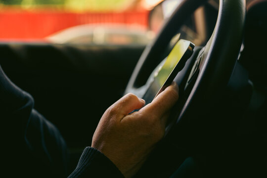 Driver using smartphone while holding steering wheel inside car, distracted driving concept