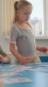 Two young girls engaged in a focused indoor activity at a table, sorting through illustrated cards in a bright room with window blinds and blue surfaces