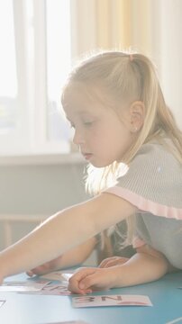 Two young girls with blonde hair engaging in an educational activity at a blue table, sorting or playing with letter cards in a warmly lit room with large windows and beige curtains