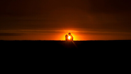 Naklejka premium Silhouette of a couple standing close together against a vibrant sunset, with the sun setting on the horizon, creating a warm glow around them