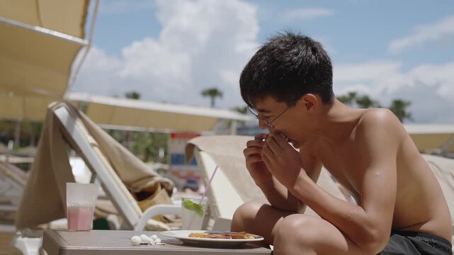Young teenager enjoying fast food snack near resort swimming pool on sunny tropical holiday. Young boy satisfy hunger with tasty lunch on sea peer during summer holidays.