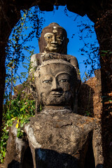 Fototapeta premium Ancient stone Buddha statues in ruined temple complex, Myanmar. Weathered historical stone Buddha sculptures surrounded by green plants and framed by an old archway under blue sky in Mrauk U, Burma